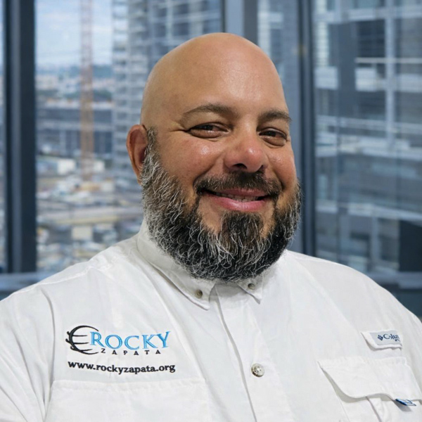 Rocky Zapata, President of Rocky Zapata Safety Consultants, smiling in a branded shirt, with a cityscape background, representing expertise in safety management and consulting services.