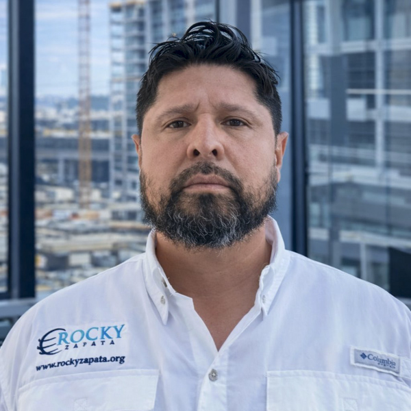 Rocky Zapata, President of Rocky Zapata Safety Consultants, wearing a branded shirt, standing in an urban setting with construction in the background, emphasizing safety management expertise.