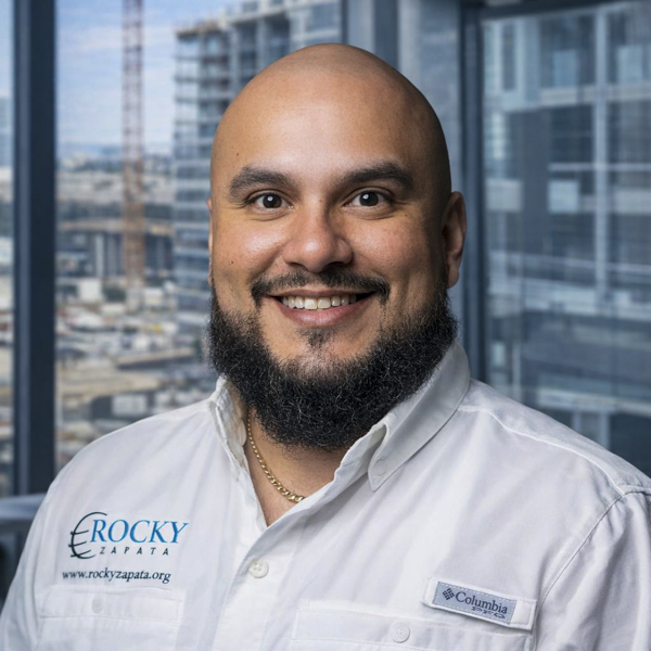 Smiling man in a Rocky Zapata Safety Consultants shirt, showcasing expertise in occupational safety, with a city skyline in the background.