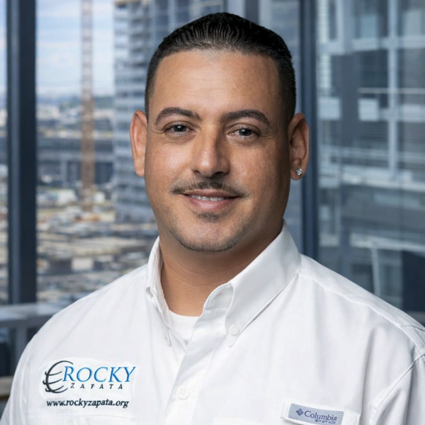 Dennis, Vice President of Rocky Zapata Safety Consultants, smiling in a white collared shirt with company logo, with a modern office background emphasizing safety consulting expertise.