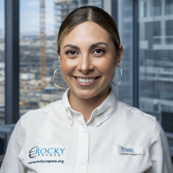 Smiling female safety consultant wearing a Rocky Zapata Safety Consultants shirt, with a city skyline in the background.