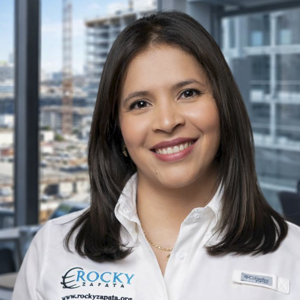 Smiling female safety consultant wearing a Rocky Zapata shirt, with a city skyline background, representing occupational safety expertise.
