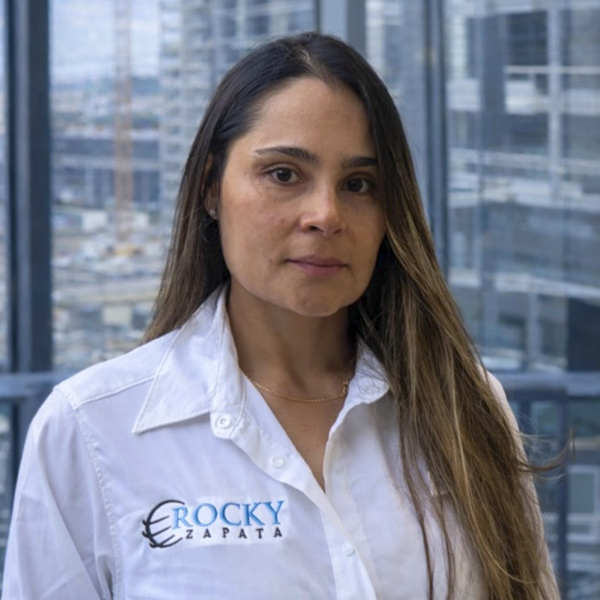 Woman in a Rocky Zapata Safety Consultants shirt, standing in an office with a cityscape background, representing the East Coast Division Safety Consultant team.
