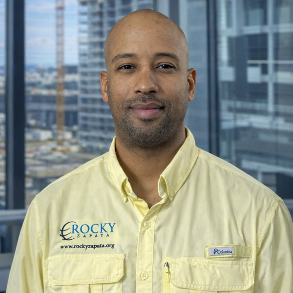 Man in a yellow safety shirt with "Rocky Zapata" branding, against a cityscape background, representing Rocky Zapata Safety Consultants and their commitment to workplace safety and training.