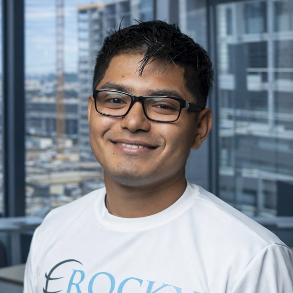 Smiling safety consultant wearing a Rocky Zapata Safety Consultants shirt, with a cityscape background.