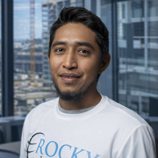 Safety consultant from Rocky Zapata Safety Consultants, wearing a branded shirt, smiling in an office setting with cityscape background.