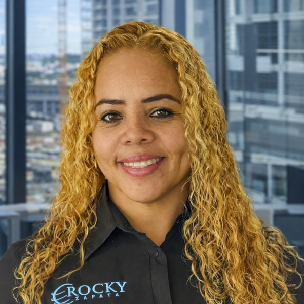 Woman with curly blonde hair wearing a black shirt with "Rocky Zapata" logo, smiling in a modern office setting, representing the team of Rocky Zapata Safety Consultants.
