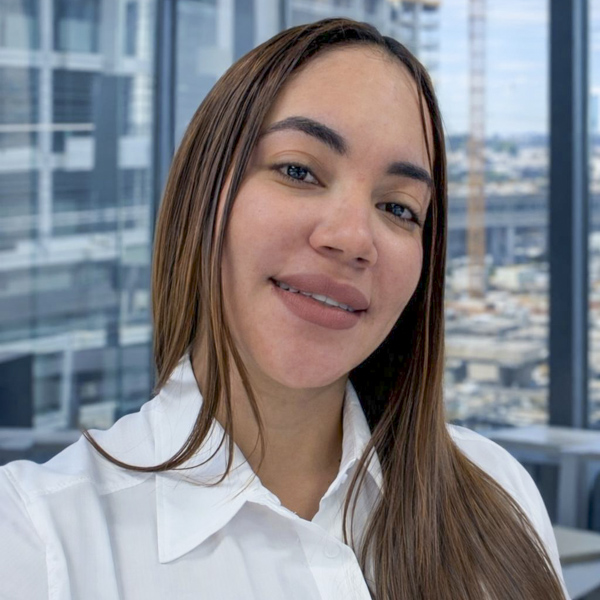 Woman smiling in a professional setting, wearing a white shirt, with a cityscape visible through large windows, representing the Rocky Zapata Safety Consultants team.