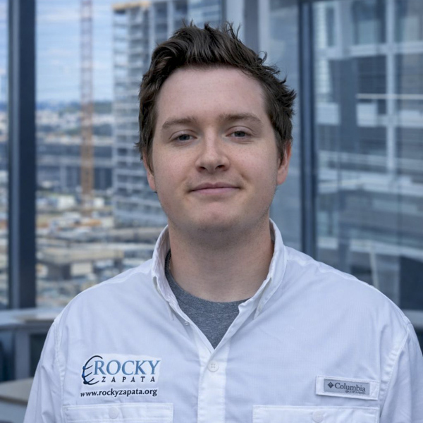 Young male safety consultant wearing a Rocky Zapata logo shirt, standing in an office setting with a cityscape view, emphasizing workplace safety expertise.
