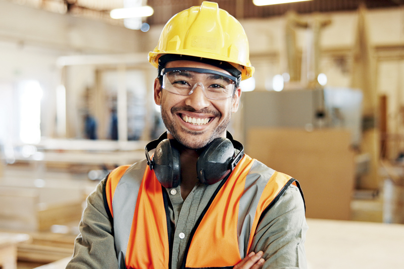Carpentry, happy and portrait of man in workshop for furniture repair, building and woodworking. Carpenter, safety gear and person with crossed arms in factory for wood, lumber and timber production Smiling construction worker wearing a hard hat, safety glasses, and ear protection, standing in a workshop environment, emphasizing workplace safety and compliance expertise.