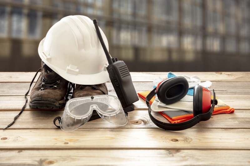 Work safety protection equipment. Industrial protective gear on Safety gear including a hard hat, work boots, safety goggles, ear protection, and a two-way radio on a wooden table, representing essential equipment for construction site safety and OSHA compliance.