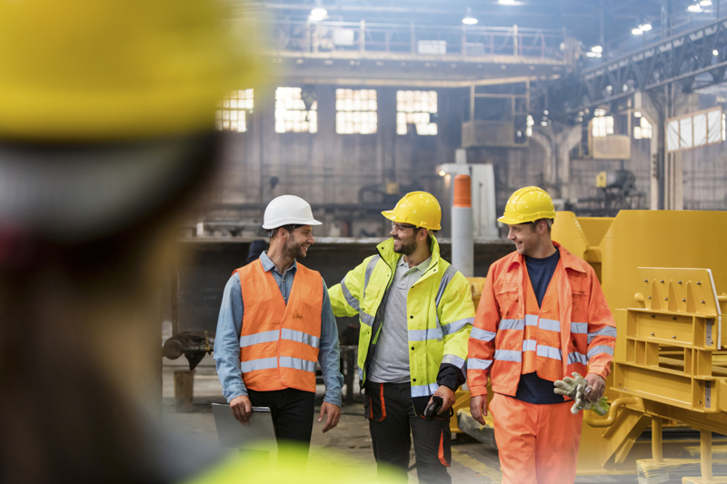 Steel workers walking and talking in factory Three safety consultants in hard hats and reflective vests discussing workplace safety in an industrial setting, emphasizing safety training and compliance in construction and general industry.